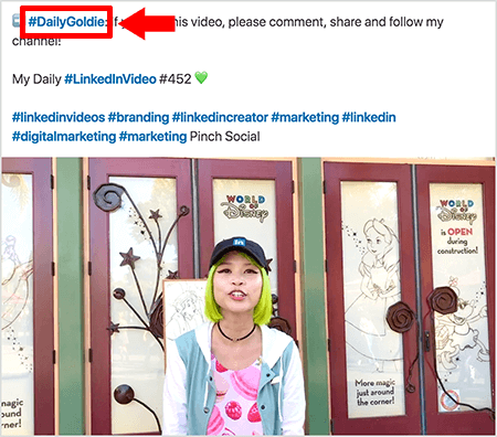 See on ekraanipilt, mis illustreerib, kuidas Goldie Chan kasutab oma LinkedIni videopostituste tekstis räsimärke. Punased tähelepanulaiendid osutavad tekstis olevale hashtagile #DailyGoldie, mis on ainulaadne tema videopostituste jaoks ja aitab tal jagamisi jälgida. Postitus sisaldab ka muid asjakohaseid räsimärke, mis aitavad inimestel tema videot leida, sealhulgas #LinkedInVideo. Videopildis seisab Goldie Disney Worldi ekraanil mõne ukse ees. Ta on roheliste juustega Aasia naine. Ta kannab musta värvi LinkedIni mütsi, musta kaelakee, roosat makaronprintidega särki ja sinimustvalget jopet.